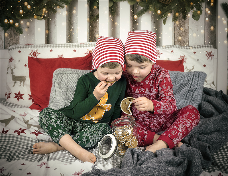 Dwóch chłopców w świątecznych piżamkach siedzi na łóżku i je pierniczki.